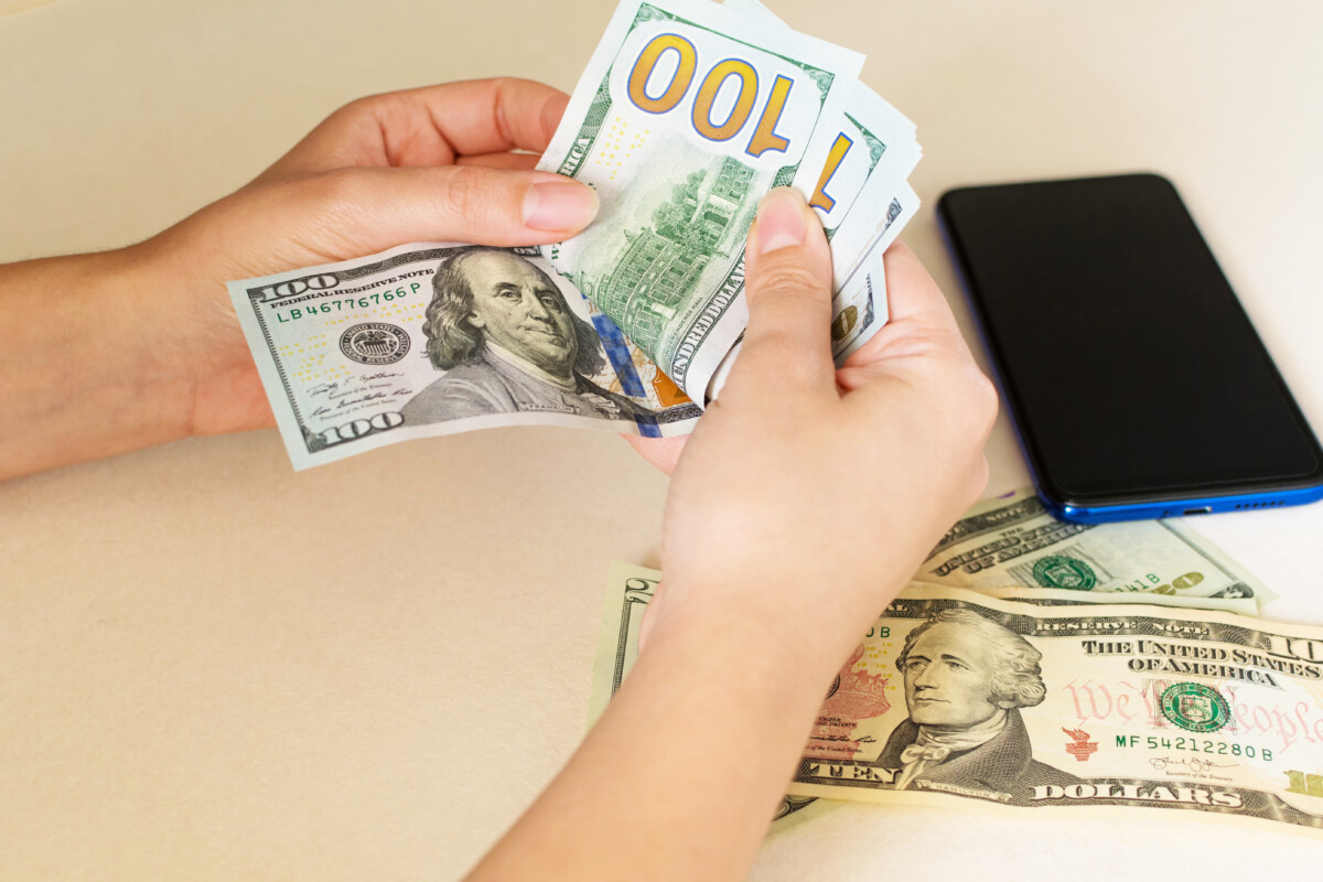 Closeup of a woman’s hands counting dollar bills, on a white desk.